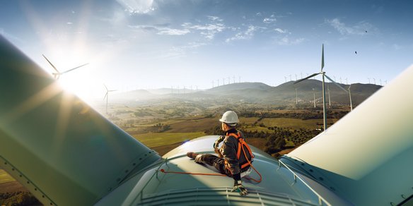 Techniker mit Sicherheitsausrüstung sitzt auf einer Windkraftanlage mit Blick auf ein Tal und weitere Windräder.