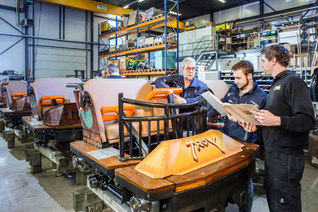  In einer Werkhalle stehen mehrere Wagen einer Achterbahn auf Holzblöcken. Die Wagen sind mit beigen Sitzen und orangefarbenen Haltegriffen ausgestattet. Im Hintergrund sind Regale mit verschiedenen Materialien und Werkzeugen. Drei Personen stehen um einen der Wagen herum, eine Person hält ein Handbuch oder eine Anleitung in den Händen.