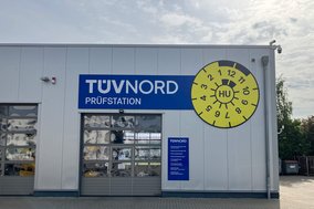 Außenansicht der TÜV NORD Station Wunstorf mit Prüfhalle, Anmeldung und große HU-Plakette an der Fassade.