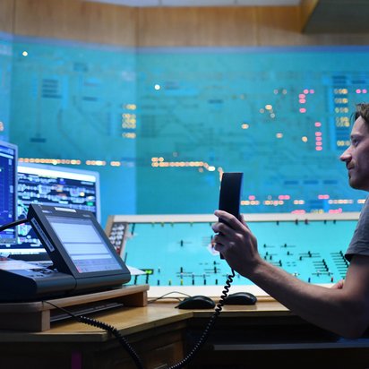 Ein Mann in grauem Shirt arbeitet in einer modernen Verkehrsleitstelle mit mehreren Monitoren und hält ein Funkgerät. Durch die große Fensterfront ist eine nächtliche Stadtlandschaft mit bunten Lichtern zu sehen.