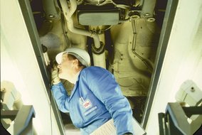 TÜV-Prüfer in blauer Arbeitskleidung inspiziert den Motorraum eines weißen Fahrzeugs von unten.