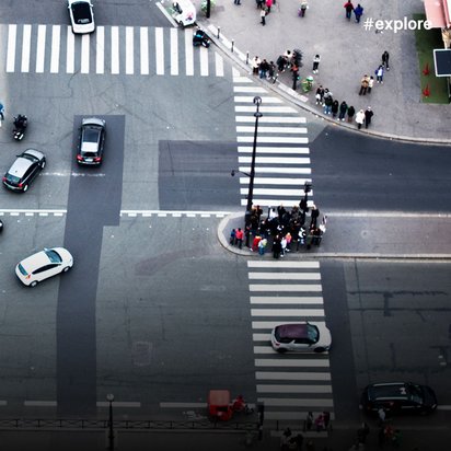 Luftaufnahme einer stark befahrenen Kreuzung mit mehreren Autos, einem Bus und Fußgängern an den Zebrastreifen. Die Kreuzung zeigt das typische Verkehrsgewimmel einer Großstadt aus der Vogelperspektive.