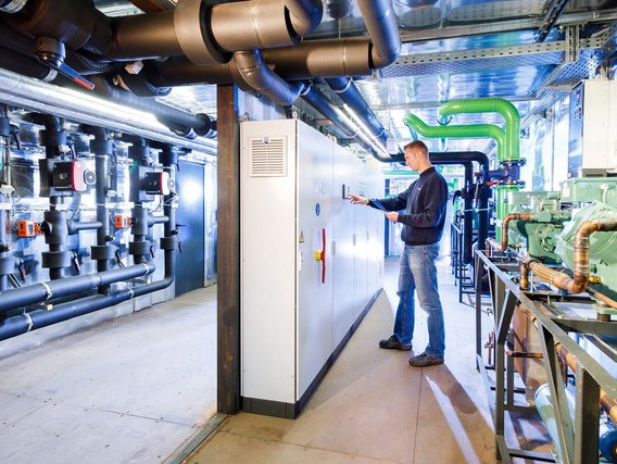 Ein Techniker in Jeans bedient ein Kontrollpanel in einem technischen Raum mit zahlreichen Rohrleitungen in verschiedenen Farben.