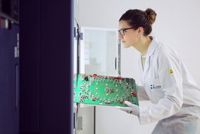  Frau mit Brille und hochgesteckten Haaren hält eine große grüne Leiterplatte mit vielen elektronischen Bauteilen. Sie trägt einen weißen Laborkittel mit ALTER TECHNOLOGY Logo.