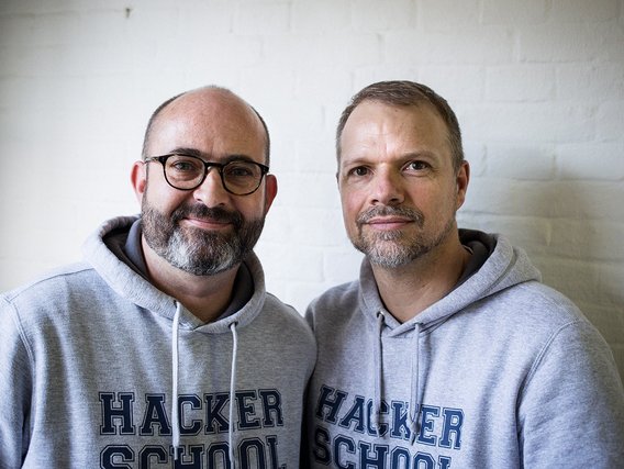 David Cummins (links) und Andreas Ollmann in grauen Kapuzenpullovern mit "HACKER SCHOOL" Aufschrift lächeln in die Kamera vor einer weißen Wand.