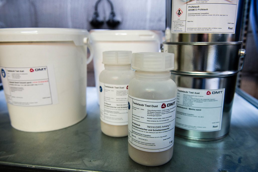 Verschiedene Teststaub-Behälter der Firma DMT stehen auf einer Laborarbeitsfläche, darunter Prüfstaub und Scheibenwaschstaub für technische Tests. Im Hintergrund sind weitere Laborgeräte und Metallbehälter zu sehen.