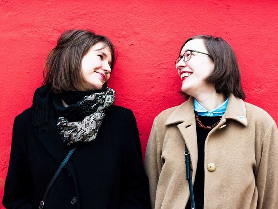 Natalie Sontopski and Julia Hoffmann stehen vor einer roten Wand, eine trägt eine schwarze Jacke, die andere einen beigen Mantel.
