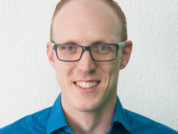 Porträtfoto: Ein lächelnder Mann mit Brille trägt ein blaues Hemd. Er hat kurze Haare und steht vor einer weißen Wand.