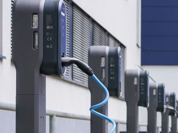 Eine Nahaufnahme einer Ladestation für Elektrofahrzeuge (EV) mit einem angeschlossenen blauen Ladekabel. Die Station ist modern und an einer dunkelgrauen Säule montiert, mit einem Gebäude mit Lüftungsschlitzen im Hintergrund.