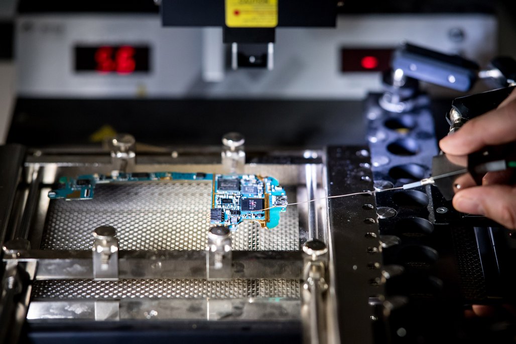 Nahaufnahme einer Mikrochip-Reparatur, wobei eine Person mit einer Pinzette einen kleinen Draht an eine Leiterplatte anbringt.