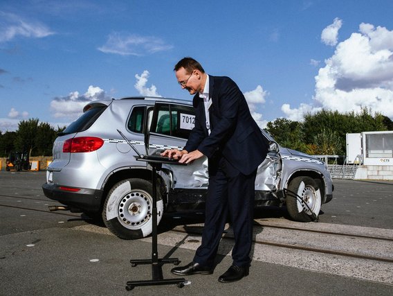 CTS-Geschäftsführer Mirko Dobberstein in Anzug bedient ein Laptop neben einem Crashtest-Fahrzeug mit Seitenschaden auf einer Testfläche.