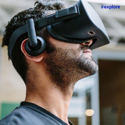 Ein Mann mit Bart trägt eine schwarze Virtual-Reality-Brille mit Kopfhörern in einem Büro oder Arbeitsbereich.