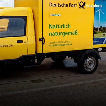Ein gelber Elektro-Lieferwagen der Deutschen Post DHL mit dem Slogan "Natürlich naturgemäß" und Windkraft-/Solarenergie-Motiven auf der Ladefläche steht an einer Ladestation.
