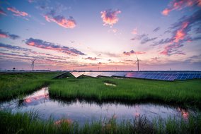 Eine Landschaft mit Solarpaneelen und Windkraftanlagen, die sich über ein grünes Feld erstrecken. Im Vordergrund ist ein kleiner Teich, der die Farben des Sonnenuntergangs am Himmel reflektiert. Der Himmel ist mit rosa und violetten Wolken bedeckt.