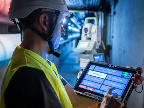 Ein Mitarbeiter mit Schutzhelm und Warnweste bedient ein Tablet vor einem Vermessungsgerät in einer industriellen Umgebung.