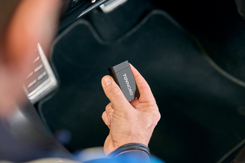 Eine Hand hält einen schwarzen TÜV Nord OBD-Adapter im Fahrzeuginnenraum vor dem OBD-Anschluss.