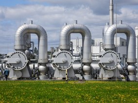 Drei große silberne Rohrleitungen mit Ventilen auf einer Industrieanlage, vermutlich für Gas oder Öl. Im Vordergrund ist eine Wiese mit gelben Blumen zu sehen.