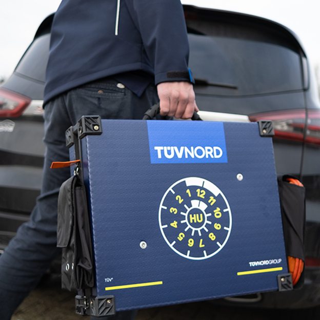 Ein schwarzes Auto von hinten, vor dem eine Person mit einem TÜV NORD gebrandeten Koffer läuft