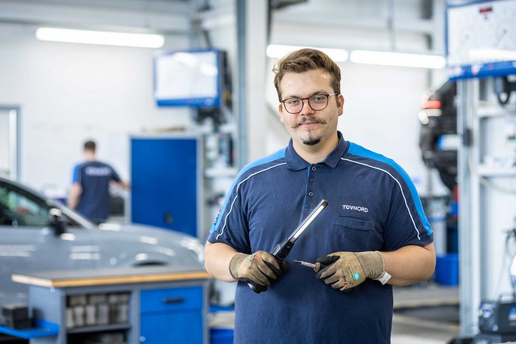 TÜV NORD Mitarbeiter mit Werkzeugen in einer Werkstatt, im Hintergrund Werkstattausrüstung, ein Auto und ein Kollege.