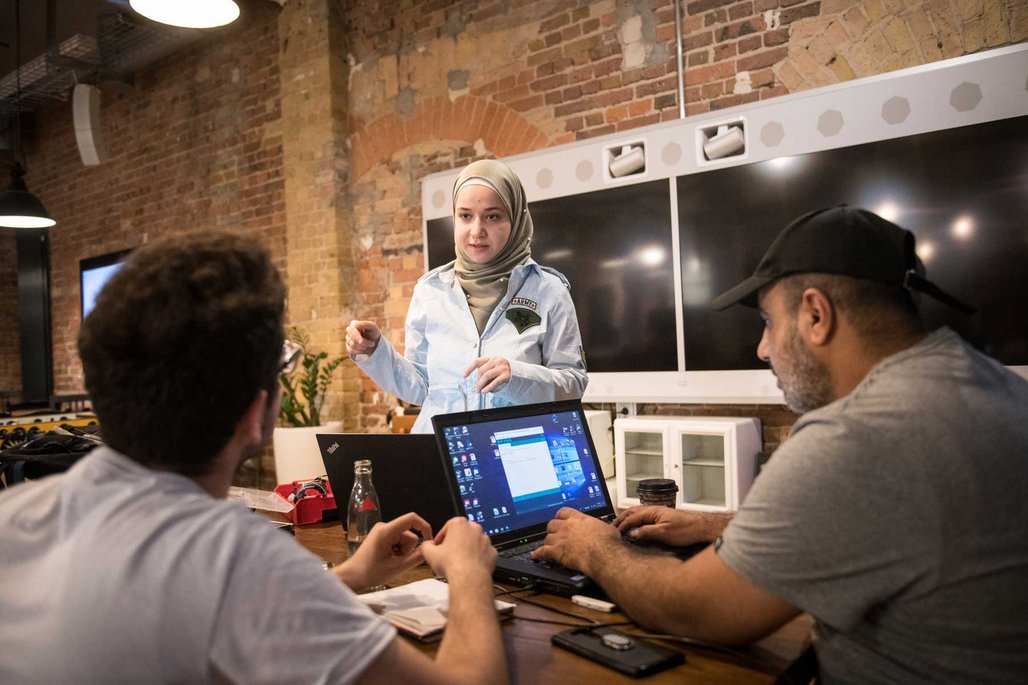 Eine Frau mit Hijab und hellblauer Jacke spricht mit zwei Personen, die an Laptops in einem Raum mit Backsteinwänden arbeiten.