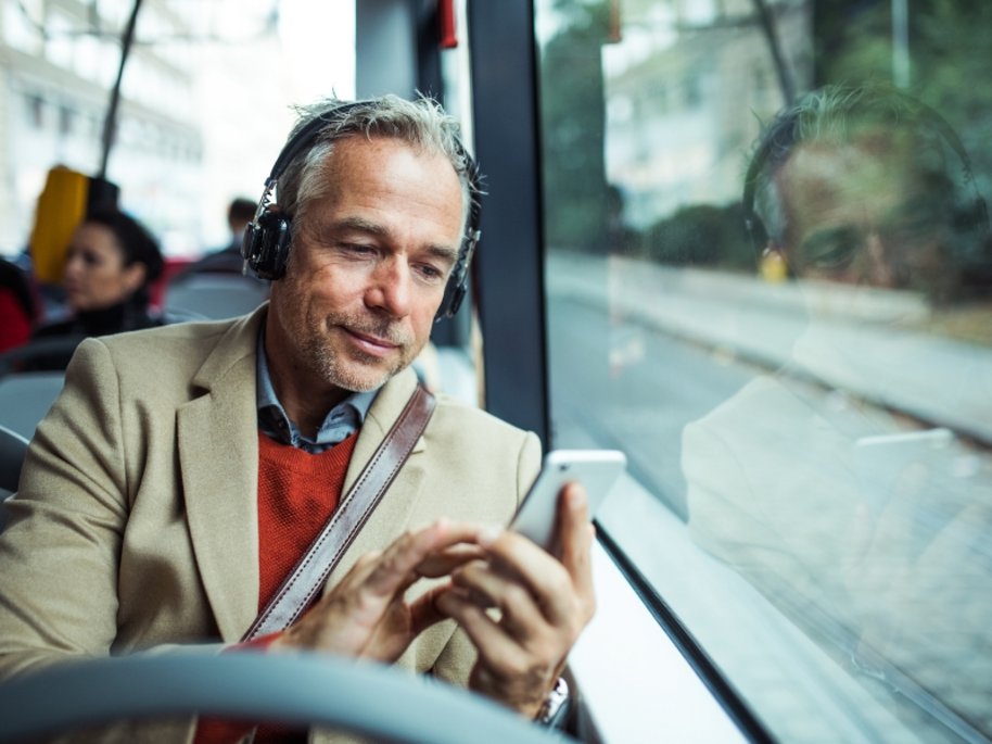 Ein älterer Mann mit Kopfhörern sitzt in öffentlichem Verkehrsmittel und schaut auf sein Smartphone.