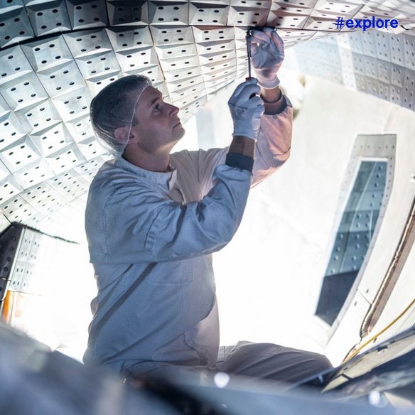 Ein Techniker in hellgrauer Schutzkleidung arbeitet im Inneren eines futuristischen, gelochten Tunnels. Er streckt die Hand nach oben und untersucht die Umgebung.