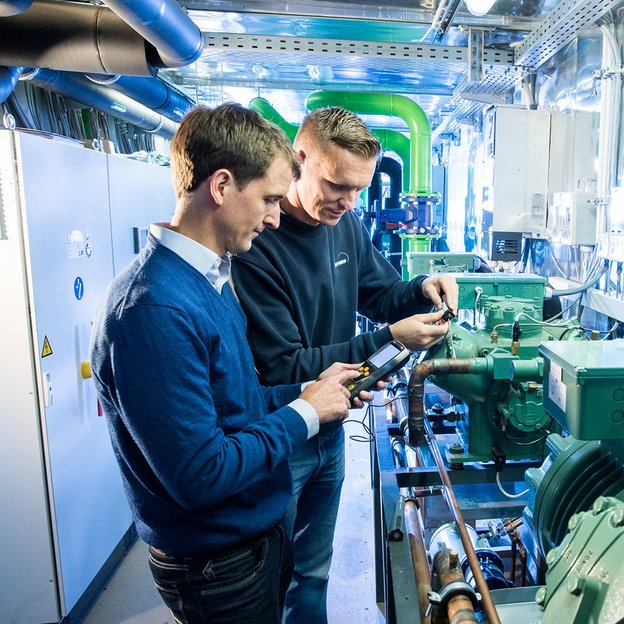Zwei Mitarbeitende stehen im Prüfbereich der Kälte-, Klima- und Lüftungstechnik bei TÜV NORD und blicken auf technische Geräte, die sie in den Händen halten.