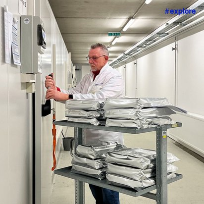 Eine Person in einem weißen Laborkittel öffnet eine Tür in einem klinischen Korridor. Daneben steht ein Rollwagen mit gestapelten silbernen Paketen.