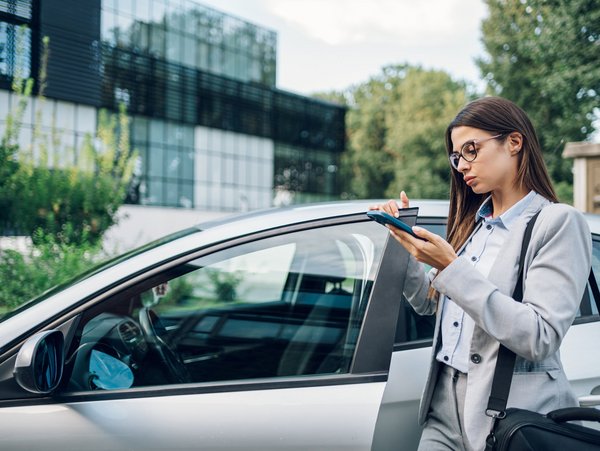 Frau im Business-Anzug mit Brille steht neben einem Auto und notiert auf einem Tablet