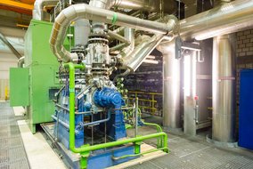 In einem industriellen Anlagenraum sind komplexe Rohrleitungen und Maschinen zu sehen. Im Vordergrund befindet sich eine große Maschine mit blauen und grünen Komponenten, umgeben von silbernen Metallrohren, die sich in verschiedene Richtungen erstrecken. Die Rohre sind teilweise mit grünen Markierungen versehen. Der Boden besteht aus einem Gitterrost, und im Hintergrund sind weitere industrielle Strukturen und Geländer zu erkennen.