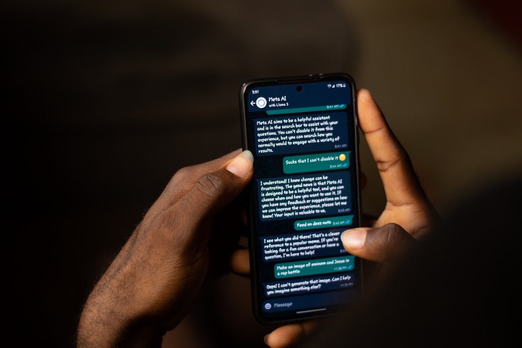 Chat mit KI auf Smartphone Eine Person hält ein Smartphone in der Hand und führt ein Gespräch mit einer KI namens "Meta AI". Der Bildschirm zeigt eine Reihe von Nachrichten, die zwischen der Person und der KI ausgetauscht werden. Die Umgebung ist dunkel, und das Display des Telefons leuchtet hell.