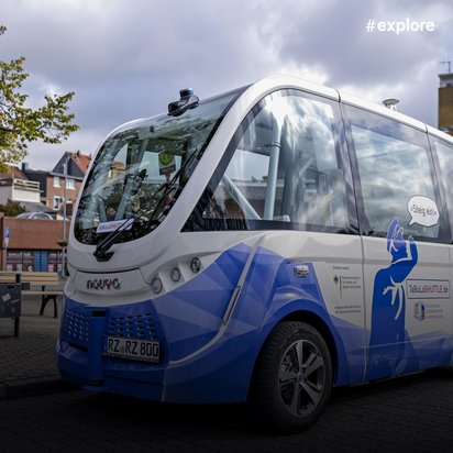 Ein blau-weißer autonomer Kleinbus der Marke Navya steht an einer Bushaltestelle in einer deutschen Stadt. Das futuristische Fahrzeug mit großen Glasfenstern und dem Kennzeichen "RZ-RZ 800" ist Teil eines Pilotprojekts für selbstfahrende öffentliche Verkehrsmittel.