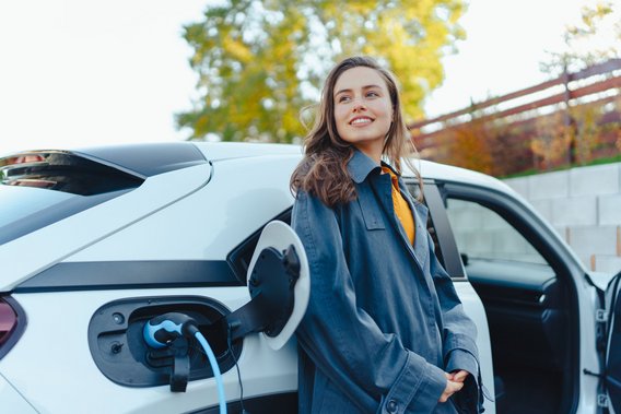 Frau steht neben einem weißen Elektroauto, das an einer Ladesäule angeschlossen ist.