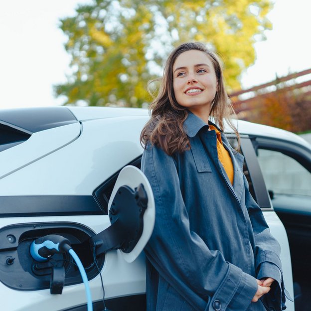 Frau steht neben einem weißen Elektroauto, das an einer Ladesäule angeschlossen ist.