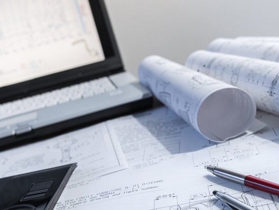 Das Bild zeigt einen Arbeitsplatz, der für technische oder ingenieurmäßige Arbeiten genutzt wird. Im Vordergrund liegen mehrere große, weiße Papierblätter mit technischen Zeichnungen und Plänen darauf. Einige dieser Pläne sind zusammengerollt. Auf den Plänen liegen zwei Kugelschreiber, einer silberfarben und einer rot-silberfarben. Links im Bild ist ein Teil eines schwarzen Zeichenbretts oder Tablets zu sehen. Im Hintergrund steht ein aufgeklappter Laptop, dessen Bildschirm ebenfalls eine technische Zeichnung zeigt. Die Szene wirkt ordentlich und professionell, als würde jemand gerade an einem technischen Projekt arbeiten oder Pläne überprüfen.