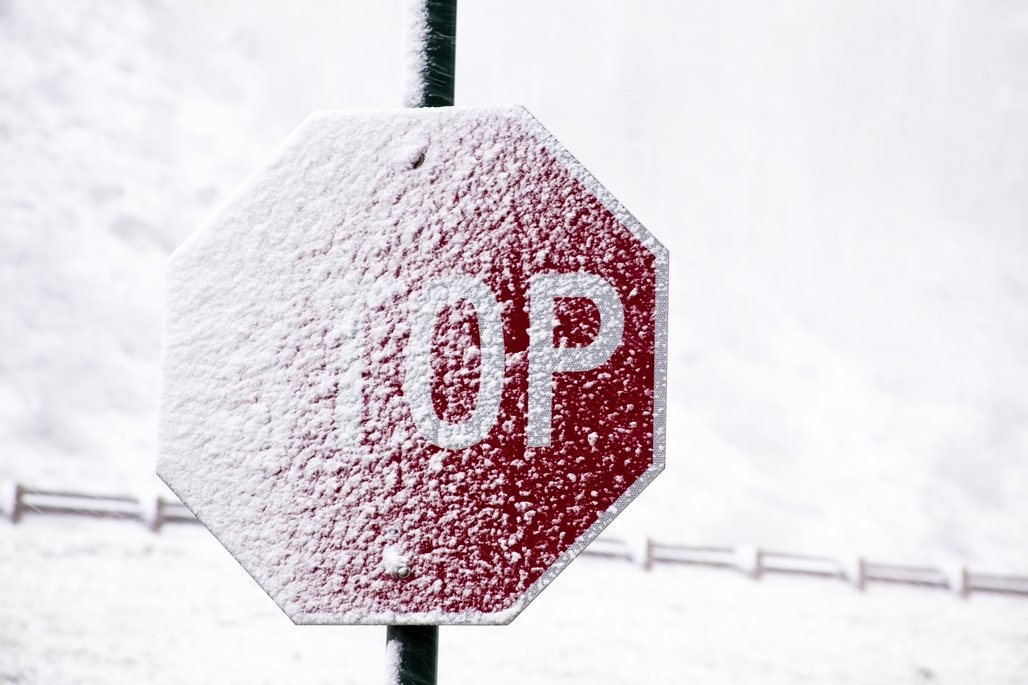 Ein verschneites rotes Stoppschild mit weißem "STOP"-Text, fast vollständig mit Schnee bedeckt, vor winterlicher Landschaft.