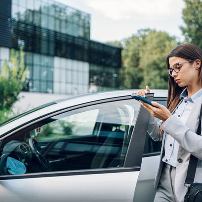 Frau im Business-Anzug mit Brille steht neben einem Auto und notiert auf einem Tablet