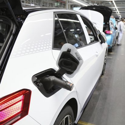 Weißes Elektroauto in einer Fertigungshalle mit geöffneter Ladeklappe und Ladestecker, Fabrikarbeiter im Hintergrund.