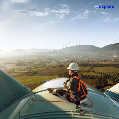 Ein Techniker mit Schutzhelm und Sicherheitsgurt sitzt auf einer Windkraftanlage mit Blick auf ein weites Tal voller Windräder bei Sonnenschein.