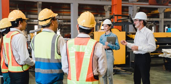Gruppe von Fachkräften mit Warnwesten und Helmen bei einer Besprechung in der Industriehalle