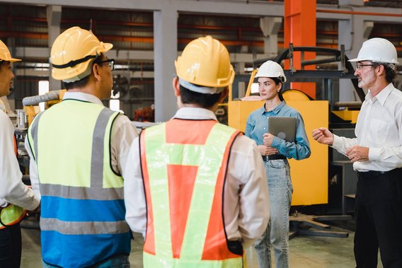 Gruppe von Fachkräften mit Warnwesten und Helmen bei einer Besprechung in der Industriehalle