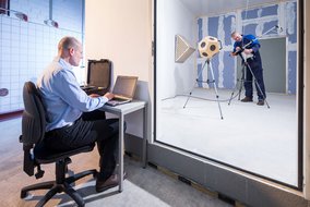 Akustikmessraum mit einer Arbeitskabine; ein Techniker arbeitet am Computer, während ein anderer im Messraum Geräte einstellt.