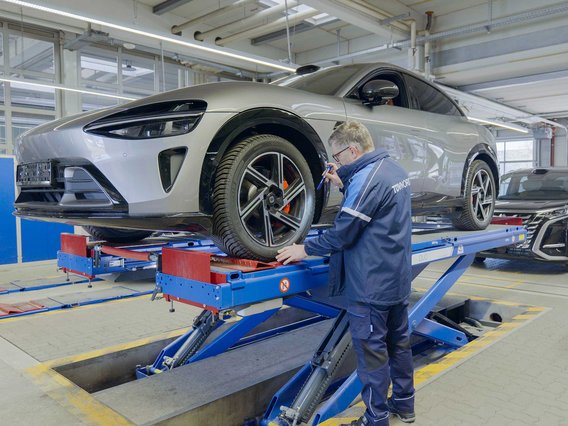 TÜV NORD Prüfer kontrolliert Fahrwerk und Bremsanlage eines aufgebockten Importfahrzeugs auf der Hebebühne, im Hintergrund ein weiteres Fahrzeug zur Begutachtung.