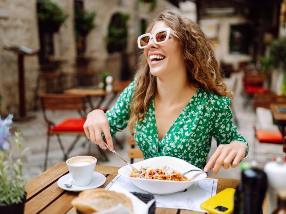 Fröhliche Frau mit lockigen Haaren und weißer Sonnenbrille im grünen Blumenkleid genießt ihr Essen auf einer sonnigen Restaurantterrasse.