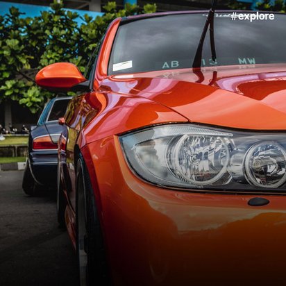 Nahaufnahme eines orangefarbenen BMW mit markanten Scheinwerfern und Kühlergrill auf einem Parkplatz. Im Hintergrund sind weitere modifizierte Fahrzeuge vor einem blauen Gebäude zu sehen.