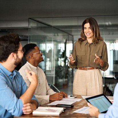 Mehrere Personen stehen und sitzen an einem Konferenztisch und besprechen gemeinsam Unterlagen in einem modernen Büro. Risikomanager (TÜV)