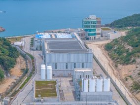 Luftaufnahme einer modernen Küsten-Industrieanlage mit grauen Gebäuden und weißen Tanks zwischen Hügeln und türkisblauem Meer.