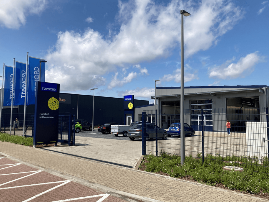 Moderne Außenansicht der TÜV NORD Station in Wolfenbüttel-Halchter mit Einfahrt, Prüfhallen und Kundenparkplätzen unter blauem Himmel.