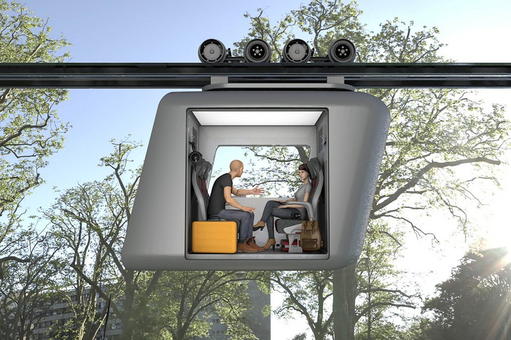 Eine futuristische Seilbahn-Gondel schwebt zwischen Bäumen, in der zwei Personen mit Gepäck sitzen und sich unterhalten. Die graue, kastenförmige Kabine hängt an einem schwarzen Schienensystem über einer Parklandschaft.