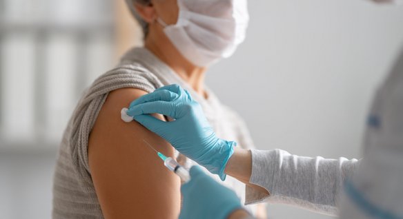 Person mit Maske erhält Injektion in den Oberarm durch medizinisches Fachpersonal mit Handschuhen.
